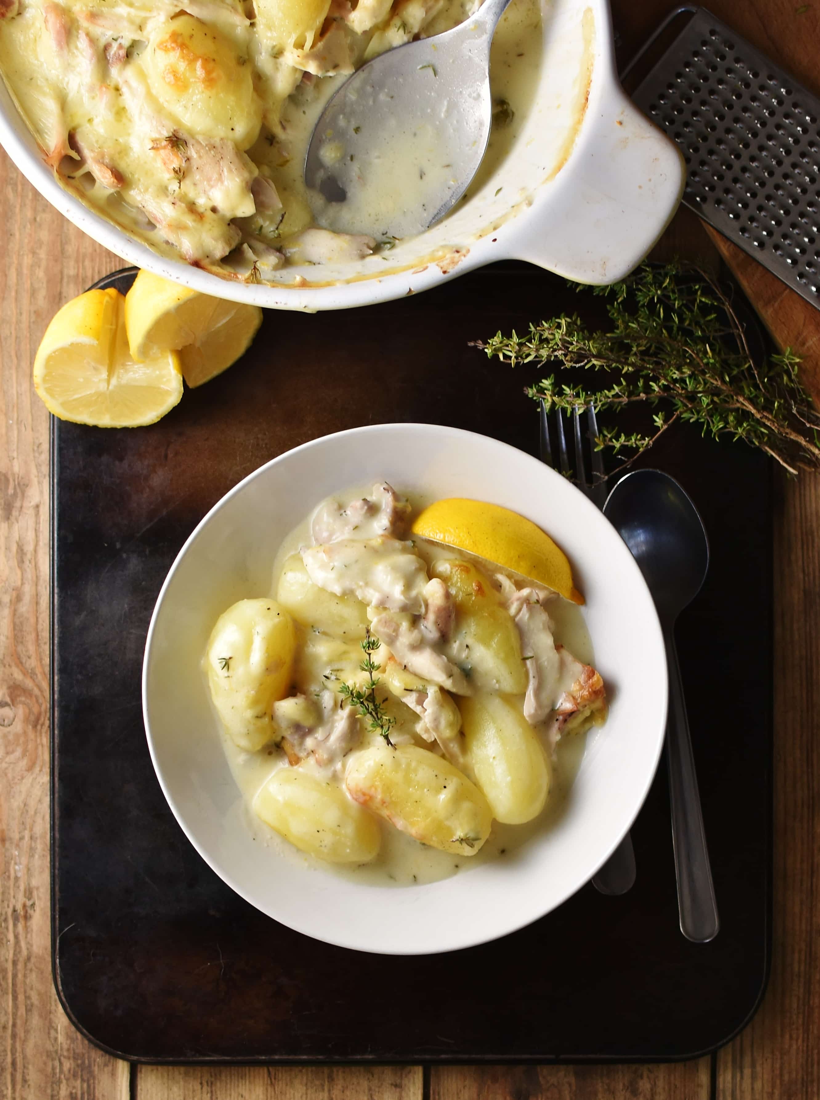 Top down view of potatoes and chicken pieces in creamy sauce with lemon wedge in white bowl, herbs and halved lemon at top and partial view of white dish.