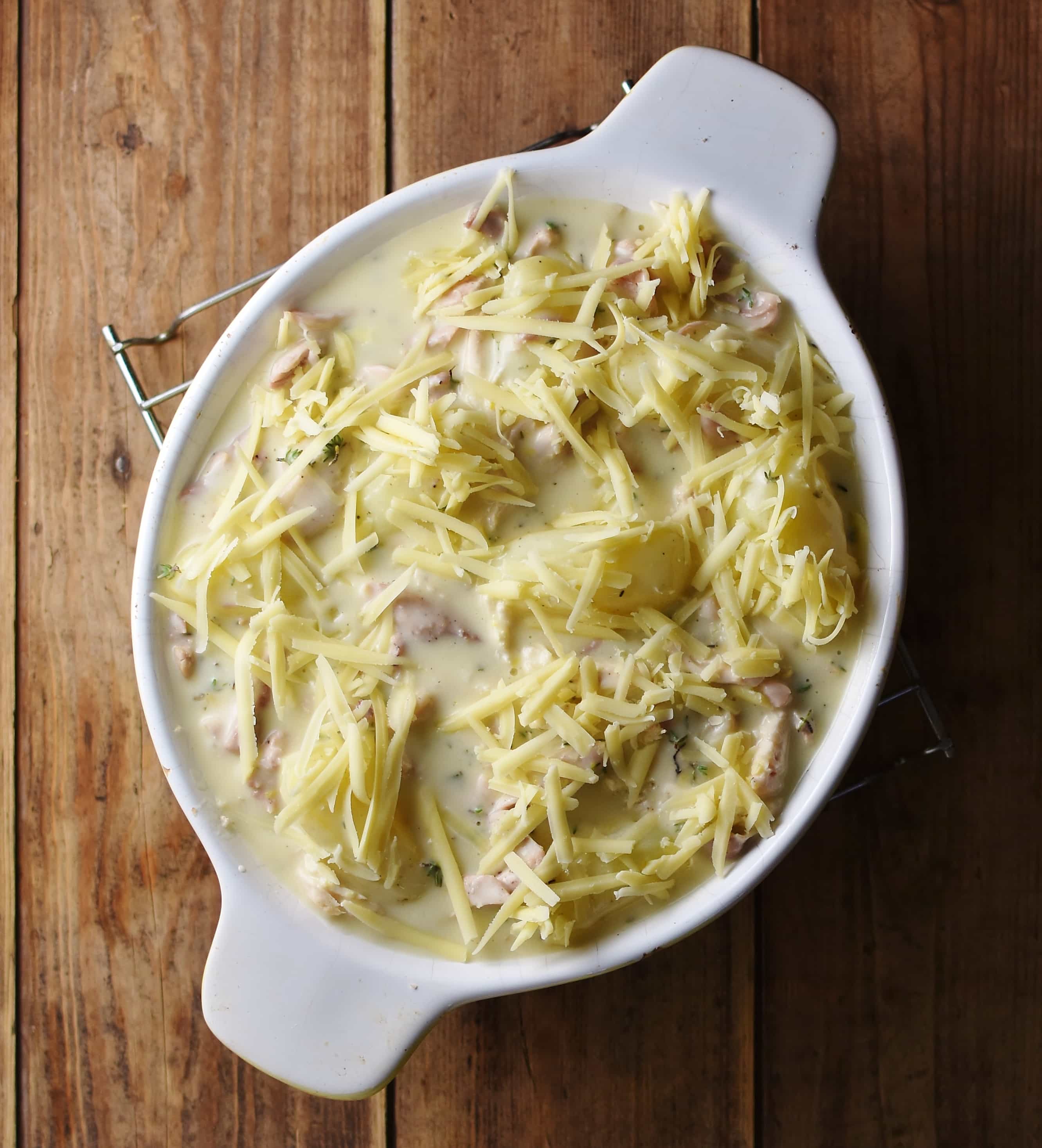 Potatoes and chicken pieces in creamy sauce with grated cheese on top in white oval dish.