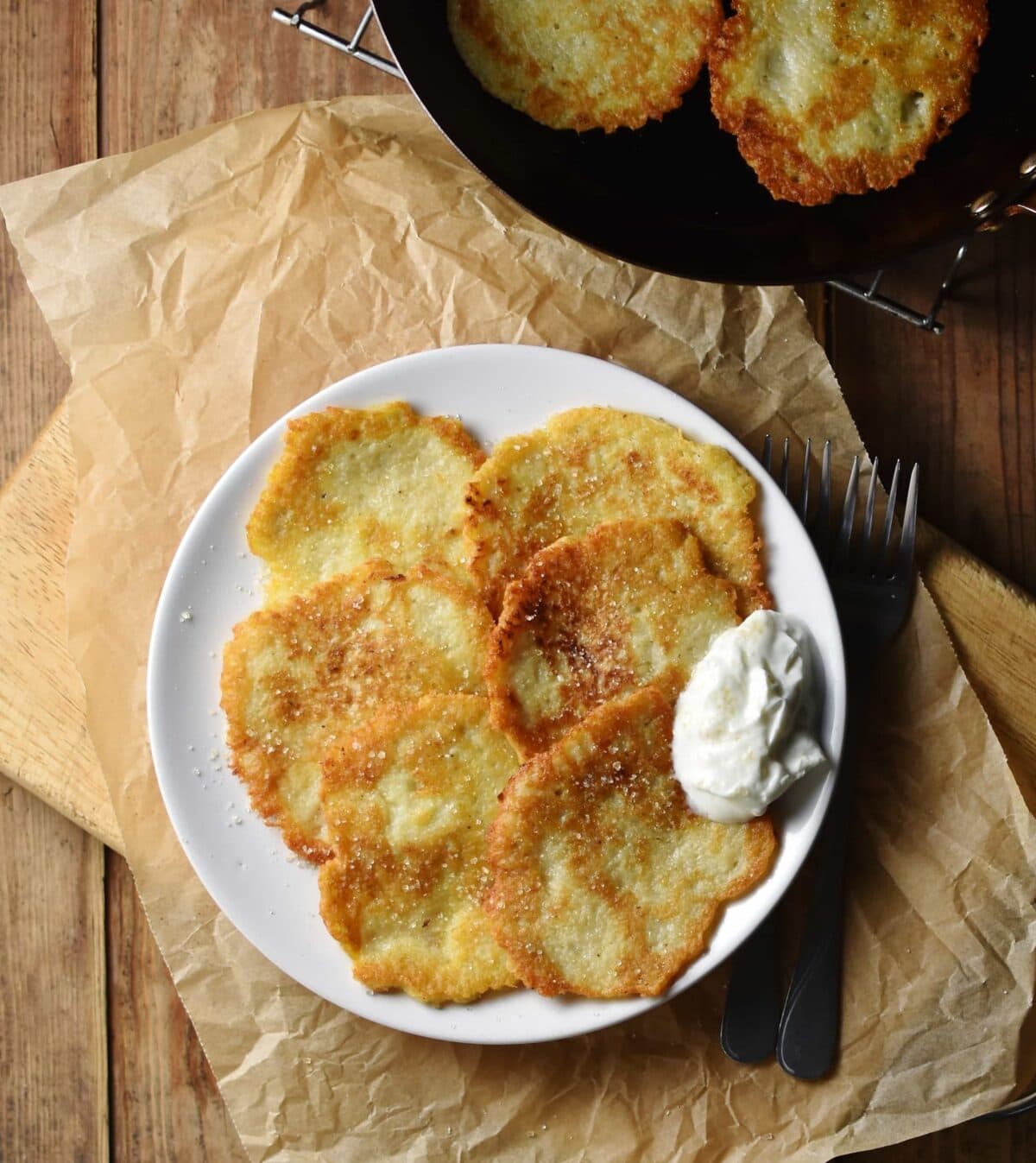 Crispy Polish potato pancakes with yogurt on white plate on top of paper, with pancakes in pan in top right corner.