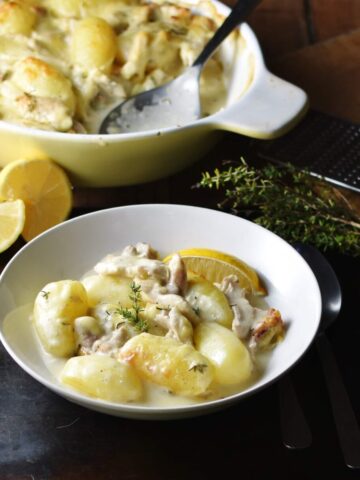 Side view of potato and chicken in creamy sauce in white bowl with yellow casserole dish and halved lemon in background.