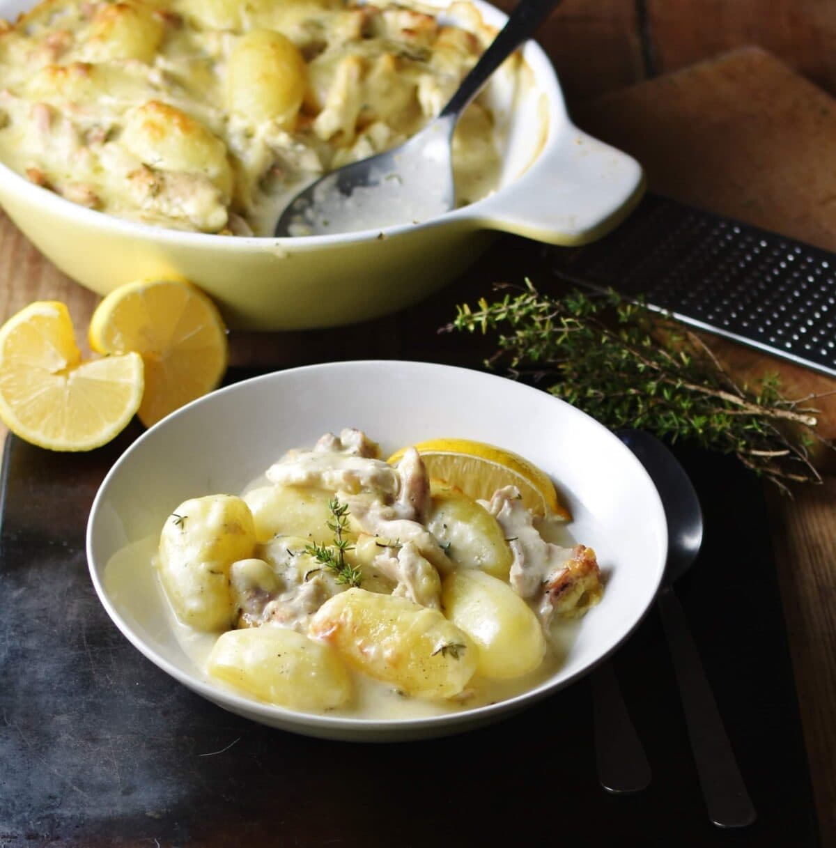 Side view of potato and chicken in creamy sauce in white bowl with yellow casserole dish and halved lemon in background.