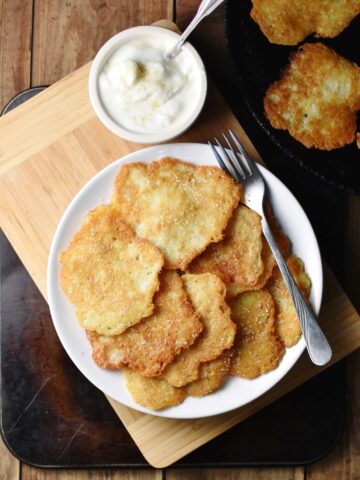 Pancakes with fork on white plate, with yogurt in small dish on top of wooden board, and with pancakes in skillet in top right corner.