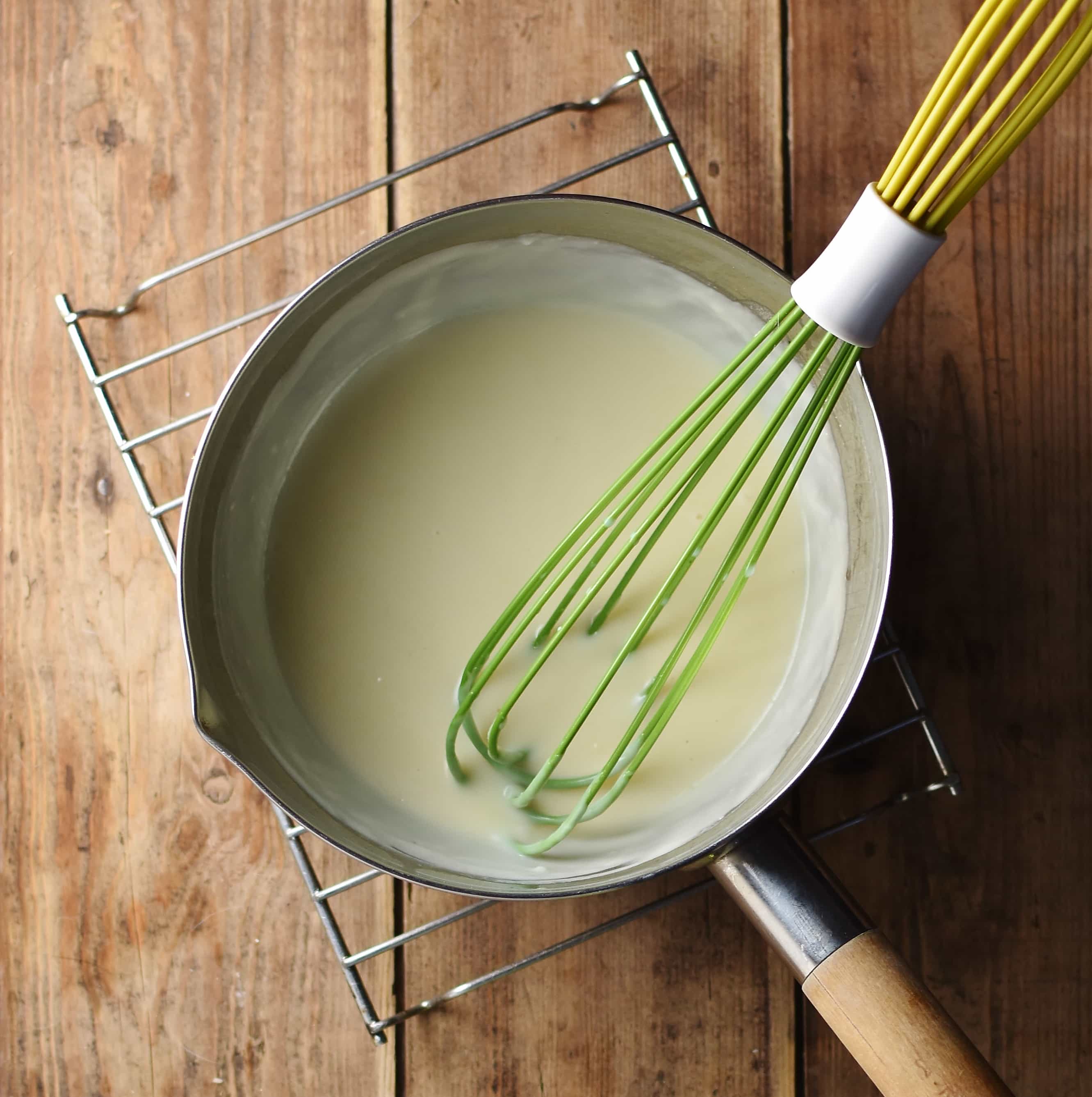 Smooth white sauce in saucepan with green whisk on top of cooling rack.