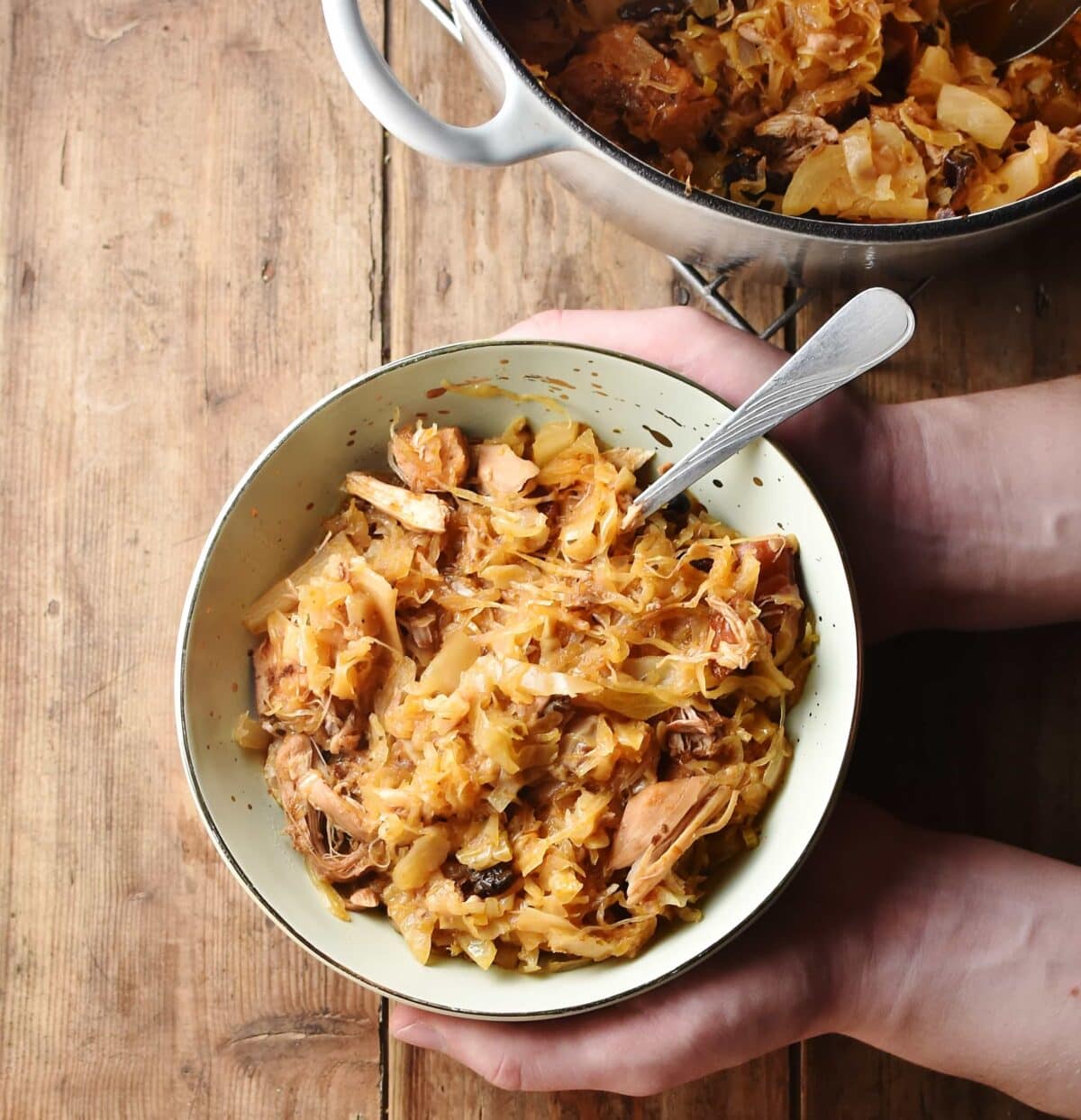 Polish bigos in green bowl with spoon held in hands, with stew in pot in background.