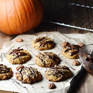 Pumpkin cookies with chocolate drizzle on top of paper with pecans, and pumpkin in background.