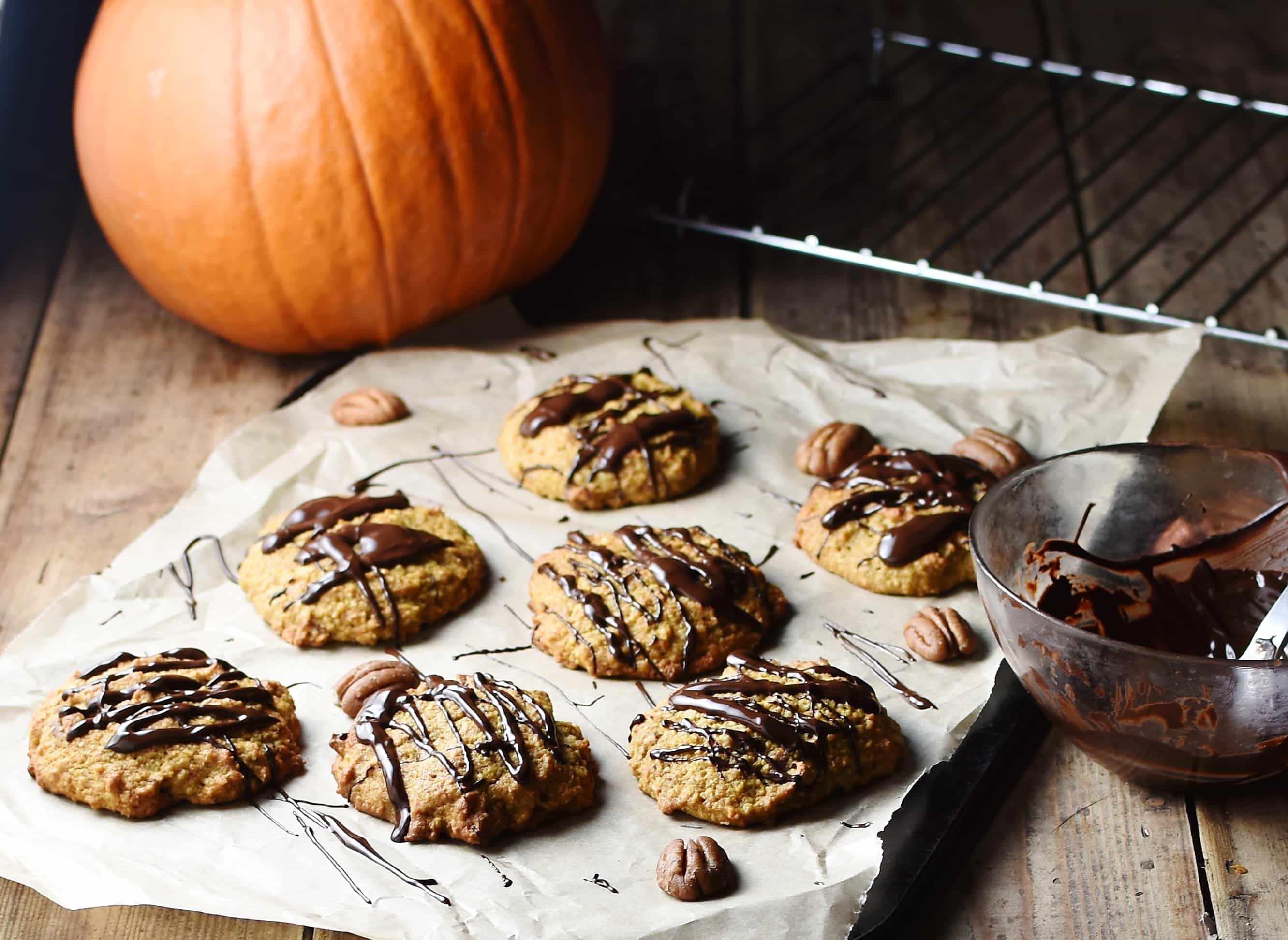 Pumpkin cookies with chocolate drizzle on top of paper with pecans, and pumpkin in background.