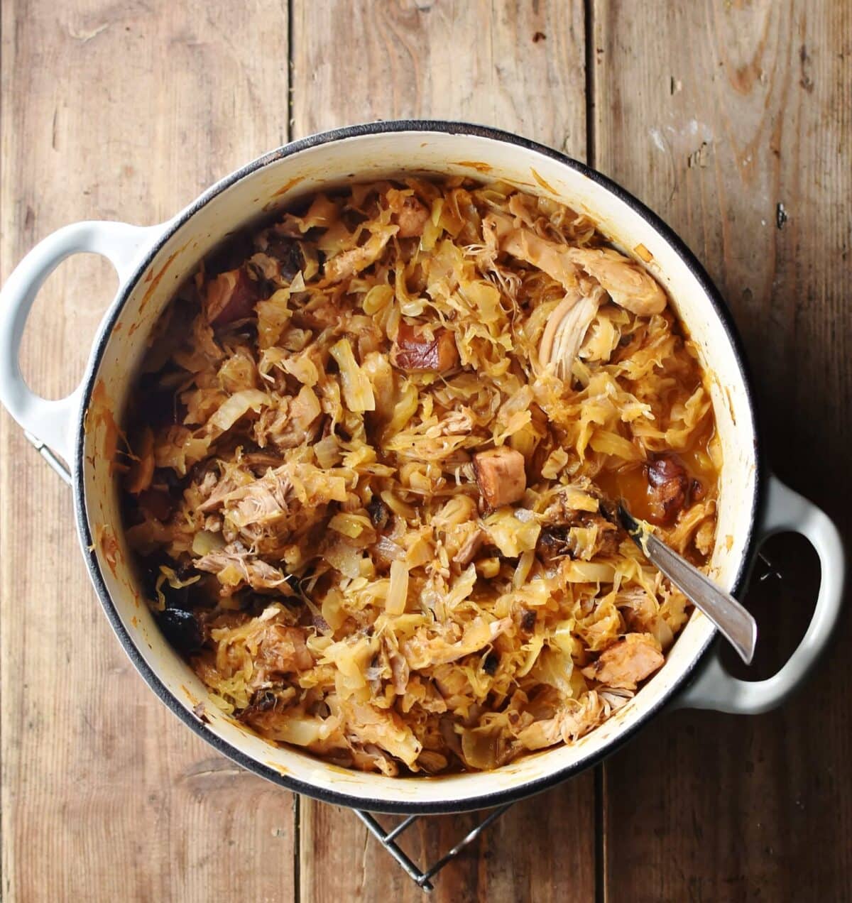 Sauerkraut with meat and mushroom pieces in large white pot with spoon.