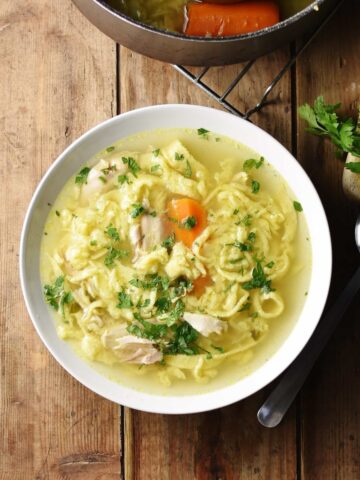 Chicken egg drop noodle soup in white bowl with spoon and parsley to right and partial view of pot at the top.
