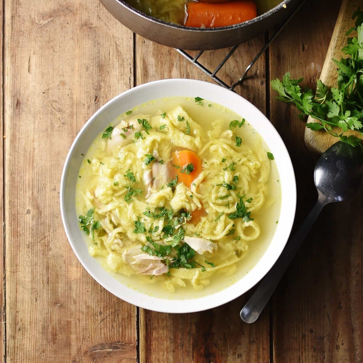 Chicken egg drop noodle soup in white bowl with spoon and parsley to right and partial view of pot at the top.