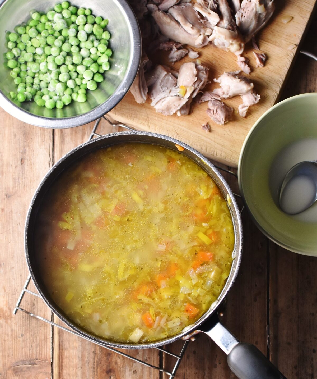 Chicken vegetable soup in large pot, with flour mixture in dish, chicken pieces on wooden board and frozen peas in bowl in background.