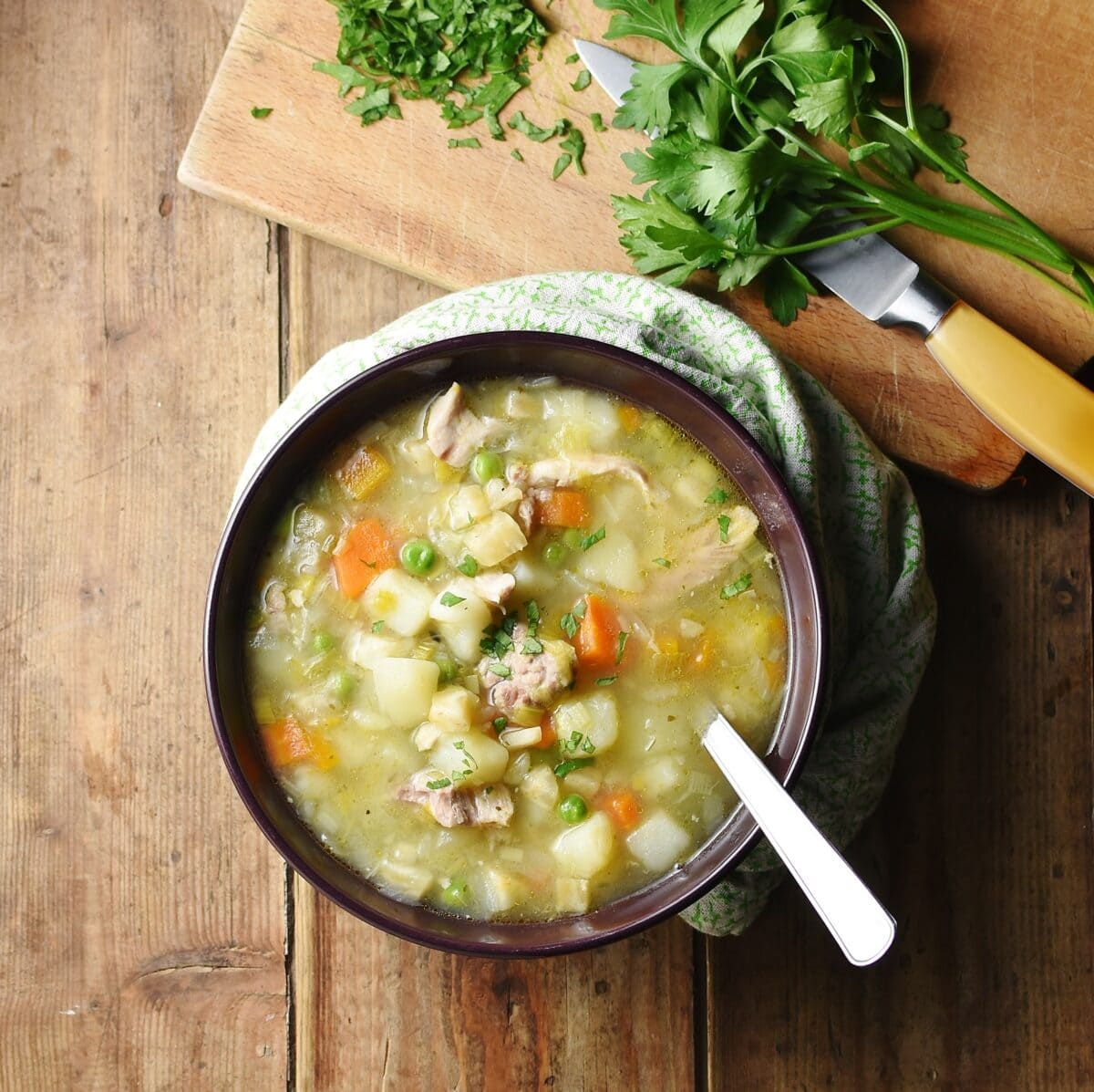 Top down view of chicken, potato and vegetable soup with spoon in purple bowl wrapped in green cloth, with fresh parsley and knife on wooden board in background.