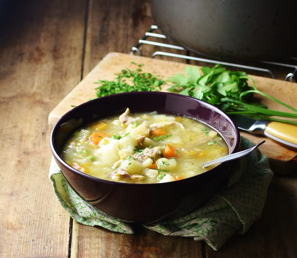 Side view of chicken potato soup with vegetables in purple bowl with spoon, herbs and pot in background.