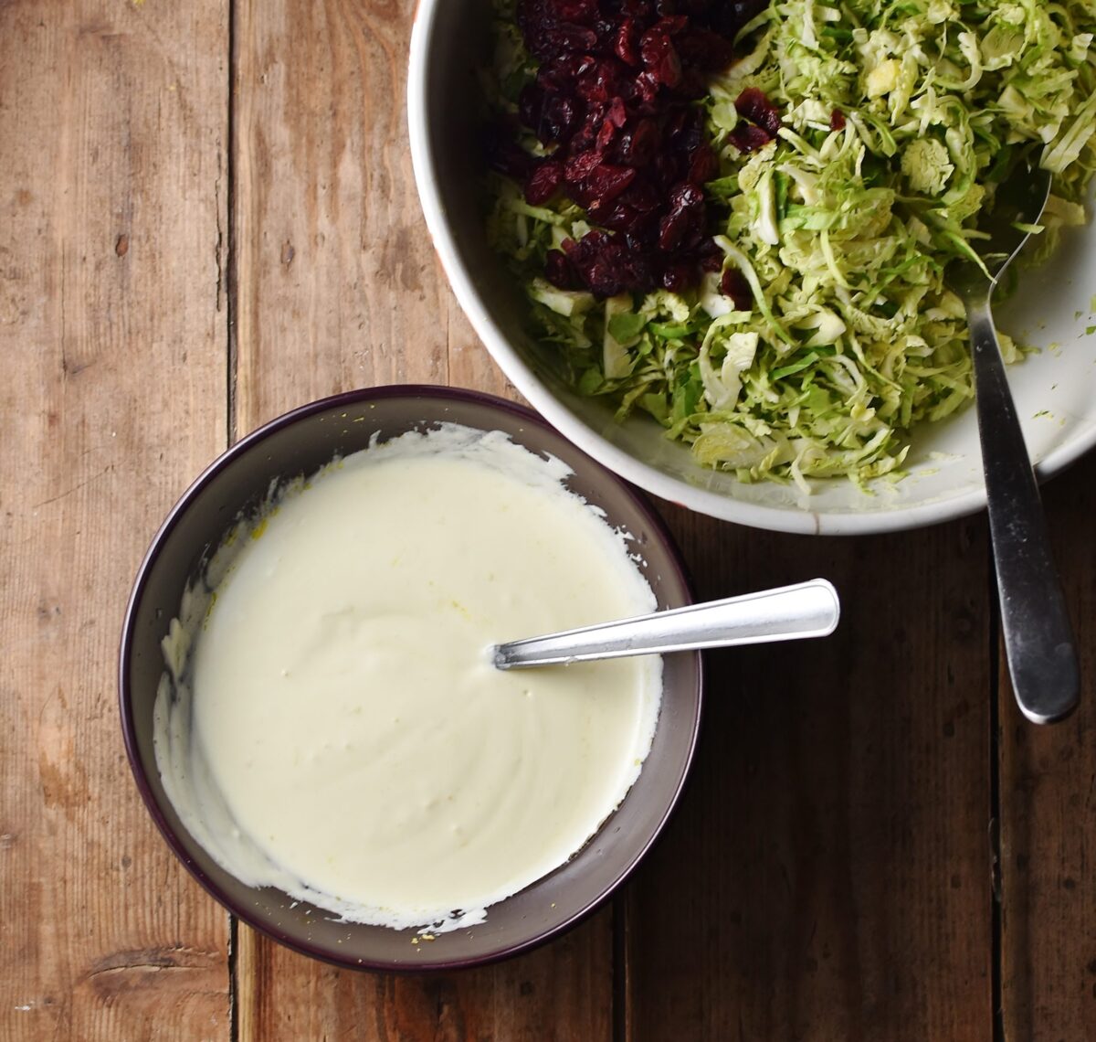 Creamy dressing with spoon in purple bowl, and shredded sprouts with cranberries and spoon in metal bowl.