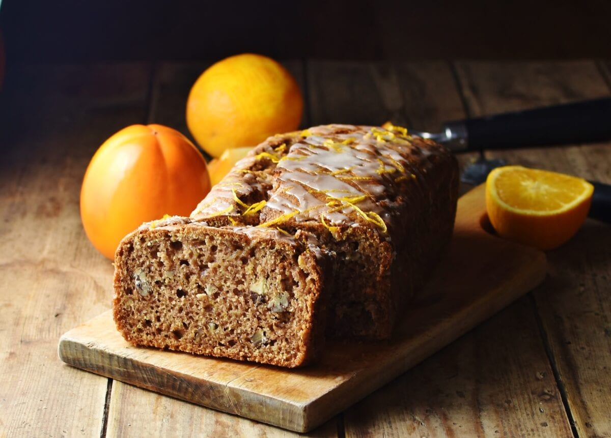 Side view of persimmon loaf on top of wooden board with persimmon and orange in background.