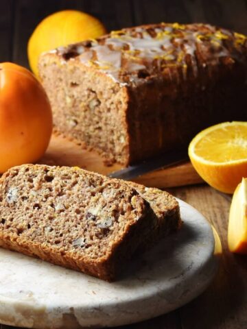 2 slices of persimmon bread on marble tray, with persimmon, orange and bread loaf in background.