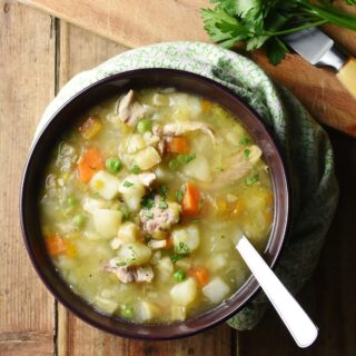 Top down view of chunky chicken, potato and carrot soup with spoon in purple bowl wrapped in green cloth, with herbs in top right.