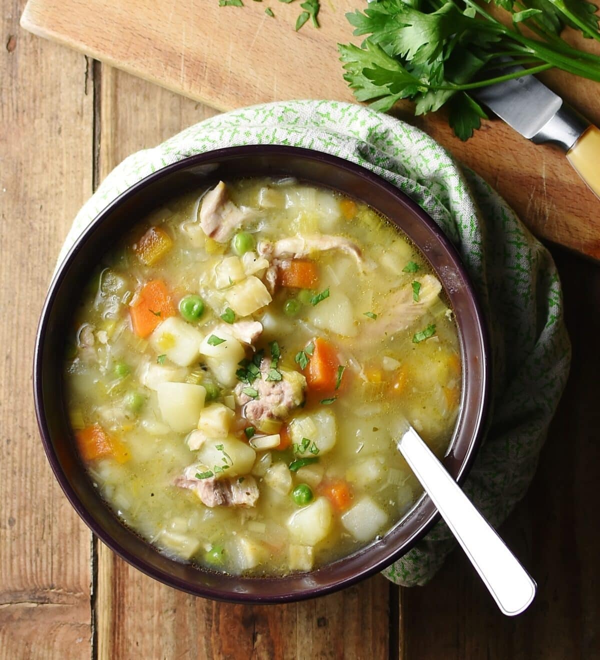 Top down view of chunky chicken, potato and carrot soup with spoon in purple bowl wrapped in green cloth, with herbs in top right.