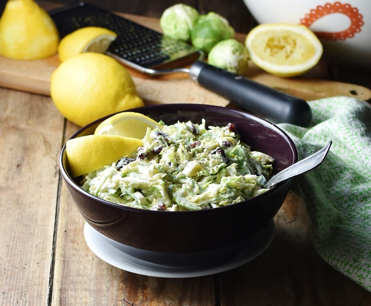 Shredded brussels sprout salad with lemon wedges and spoon in purple bowl on top of white plate, with lemons, zester and brussels sprouts in background.