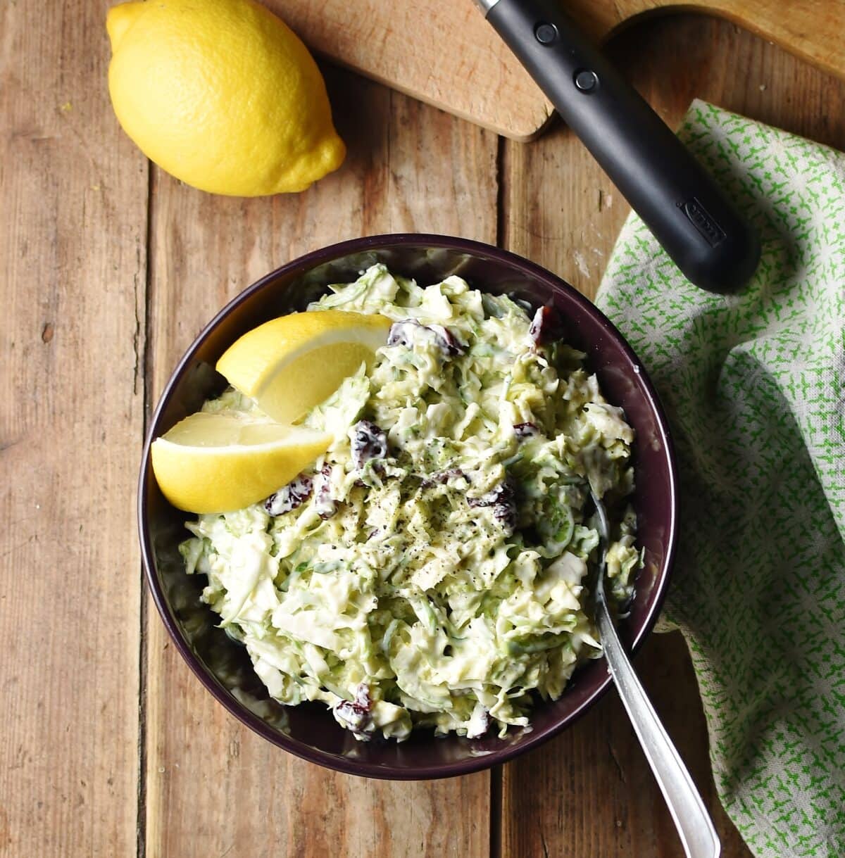 Top down view of creamy brussel sprout slaw with lemon wedges and spoon in purple bowl, with green cloth and lemon in background.