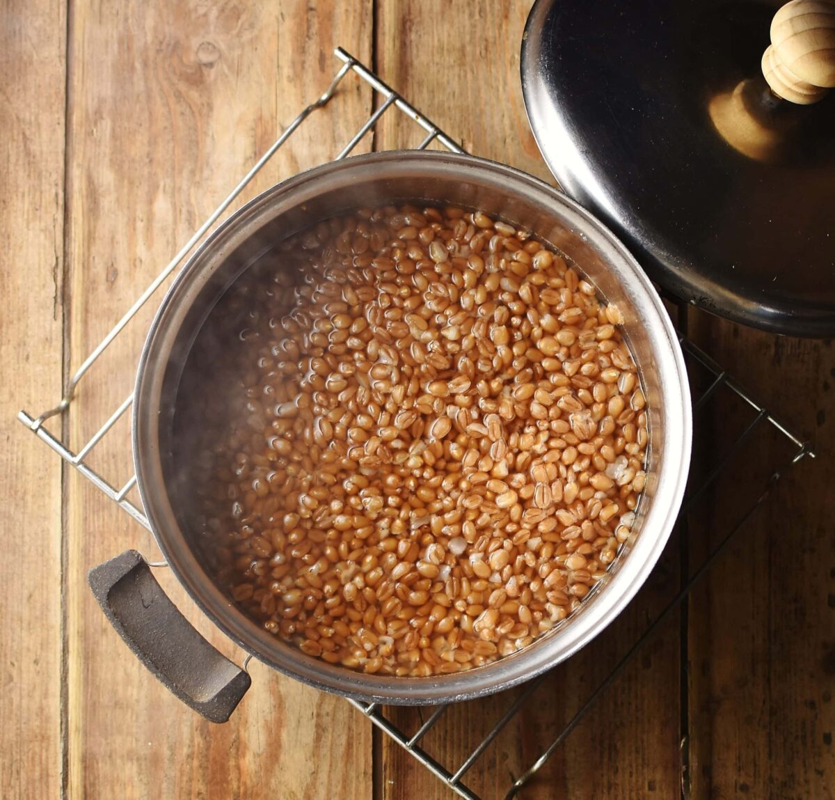 Wheat berries in water in pot with steam coming out, with lid in top right.