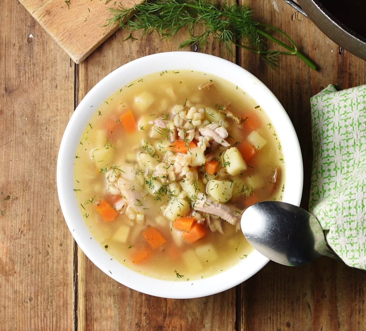 Chunky potato, carrot and barley soup in white bowl with spoon, green cloth to the right and dill at the top.