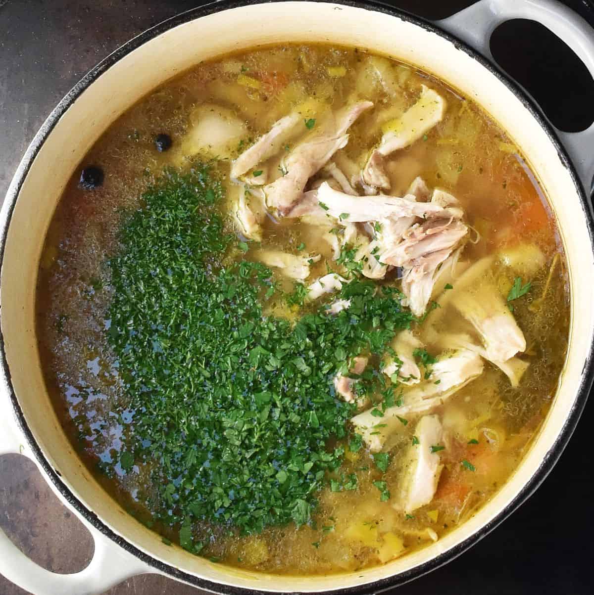 Pieces of chicken and fresh herbs in barley broth in large pot.