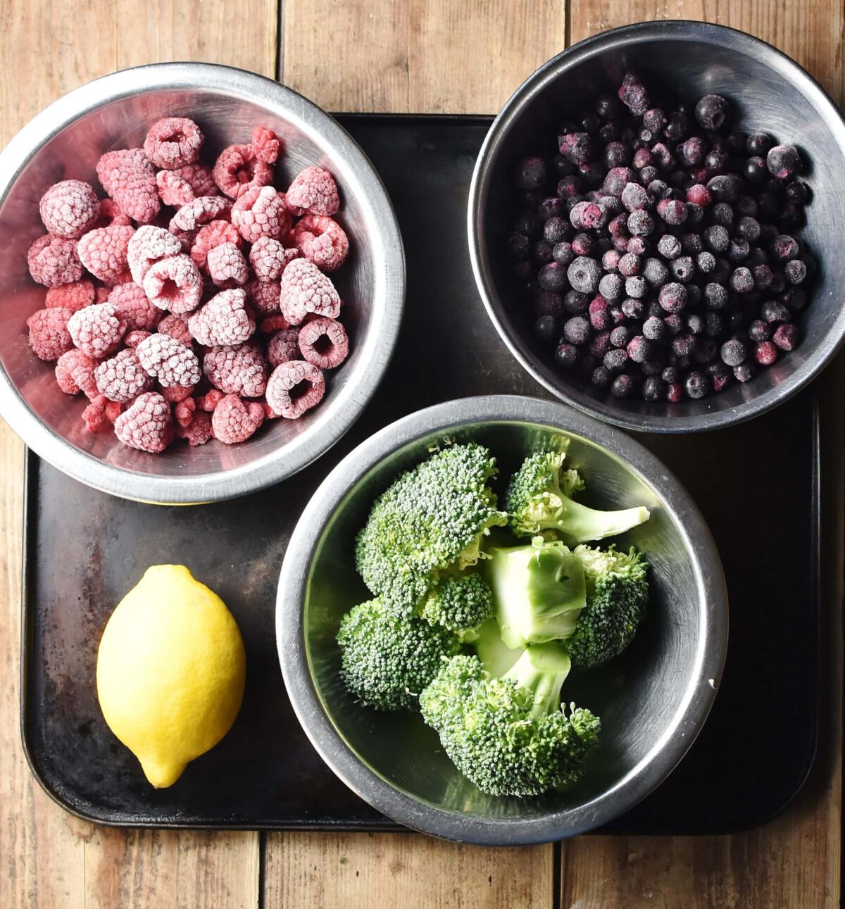 Frozen blueberries, raspberries and chopped broccoli in individual metal bowls and lemon on top of black tray.