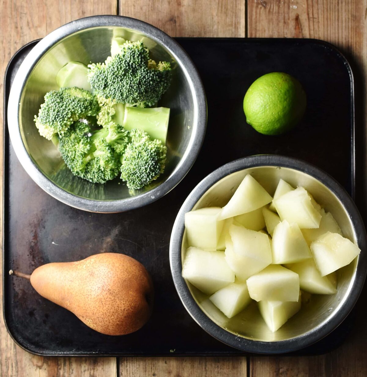 Chopped melon and broccoli in separate metal bowls, pear and lime on top of black tray.