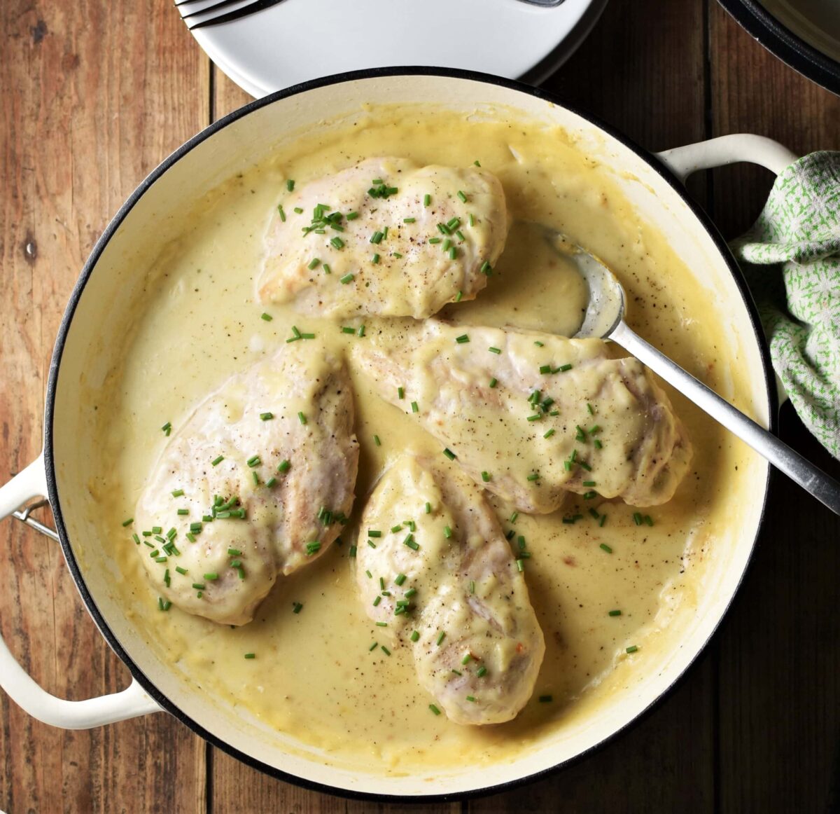 Top down view of 4 chicken breasts in white sauce with spoon in large white pan, with green cloth to the right and plates at the top.