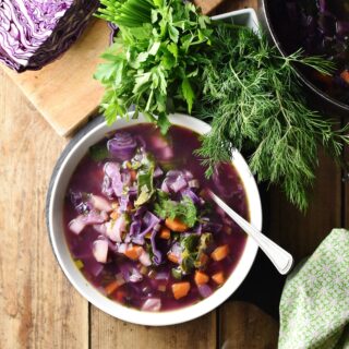 Chunky red cabbage vegetable soup in white bowl with spoon, dill, parsley, green cloth and red cabbage in background.