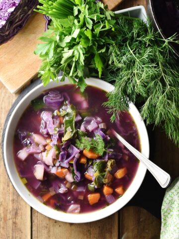 Chunky red cabbage vegetable soup in white bowl with spoon, dill, parsley, green cloth and red cabbage in background.