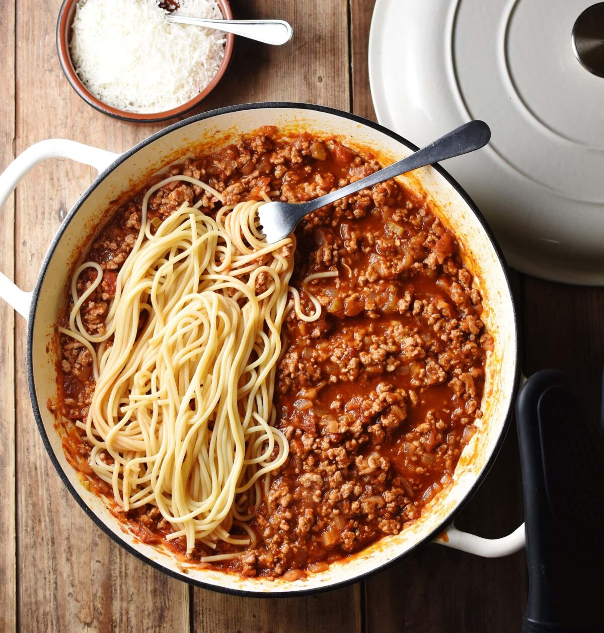 Top down view of turkey bolognese sauce with spaghetti and fork in large white shallow dish, with grated cheese in small dish and white lid in background.