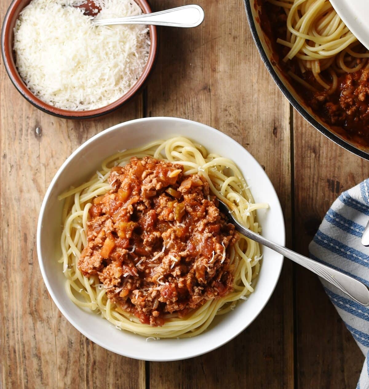 Spaghetti bolognese in white bowl with fork, grated cheese in brown dish with spoon, spaghetti in pan and stripy blue cloth in background.