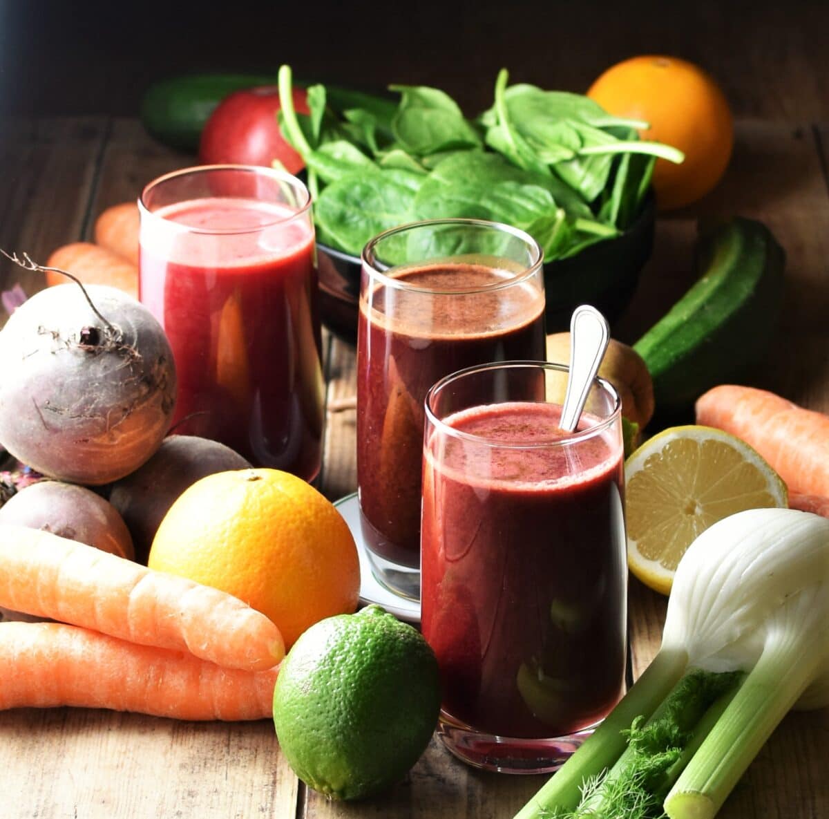 3 glasses of dark beetroot juice with fresh vegetables and fruits in background.