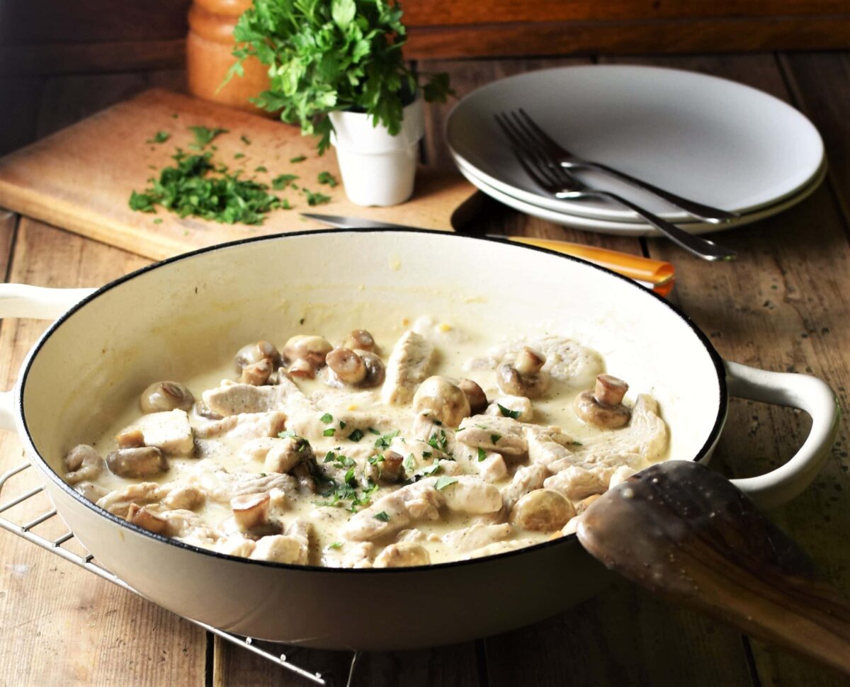 Side view of creamy turkey stroganoff with mushrooms in large white shallow pan, with herbs on top of wooden board and plates with forks in background.