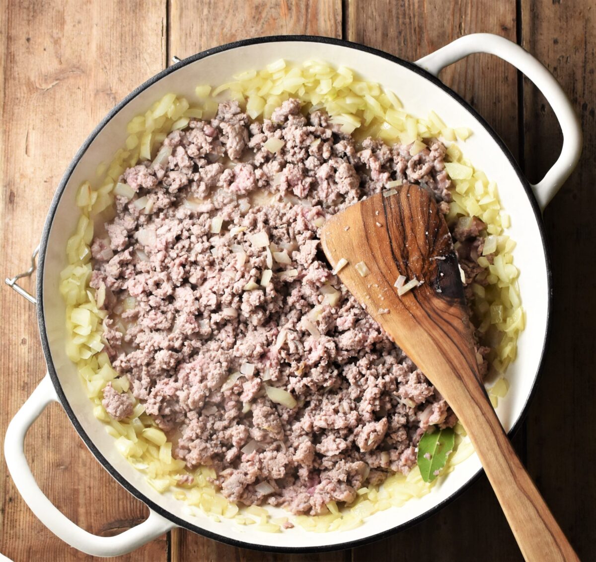 Onion and ground meat mixture in large white shallow pan with wooden spoon.