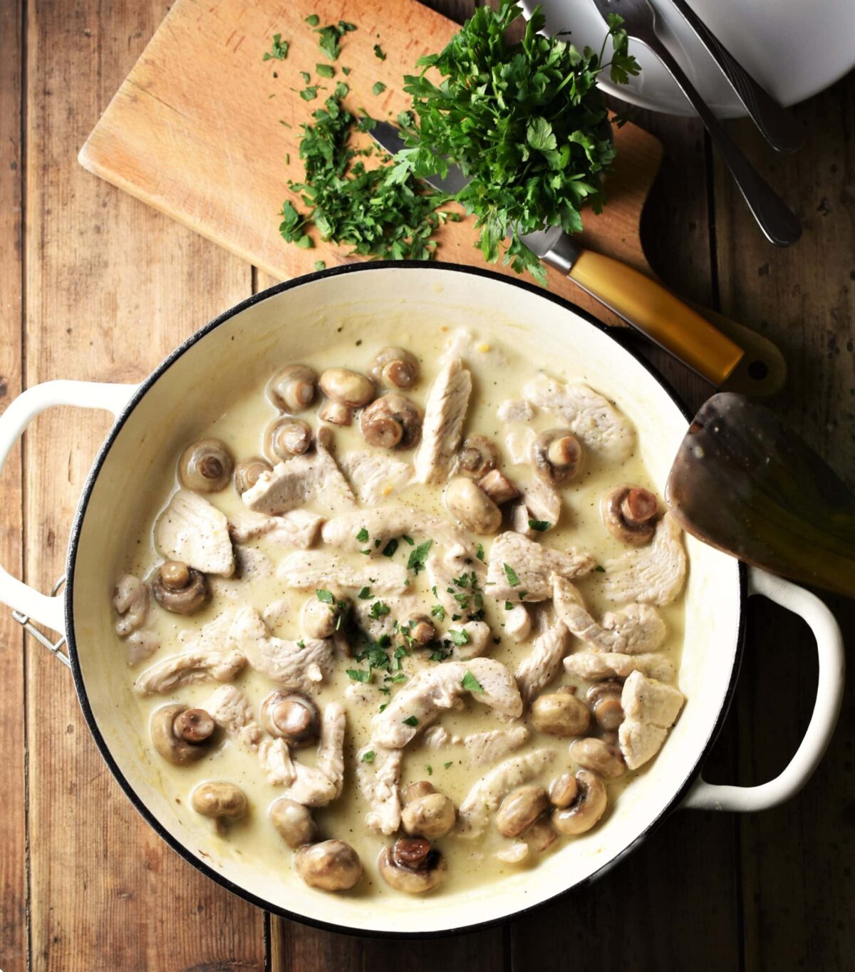Creamy turkey and mushroom in large white pan, with chopped herbs on top of wooden board with knife in background.