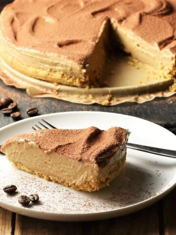 Side view of coffee cheesecake slice on white plate with fork and coffee beans, with cheesecake and spatula in background.