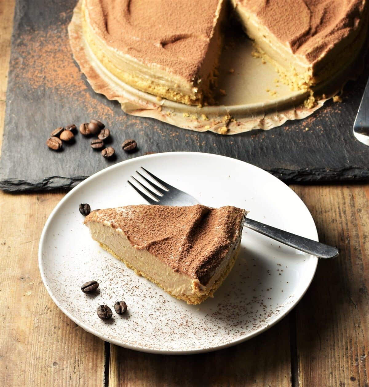 Slice of coffee cheesecake on white plate with coffee beans and fork, with cheesecake in background.