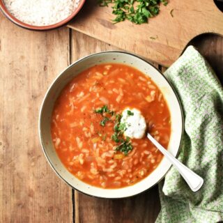 Tomato rice soup with yogurt and herbs in green bowl with spoon, green cloth the the right and rice in dish on top of wooden board with chopped herbs in background.