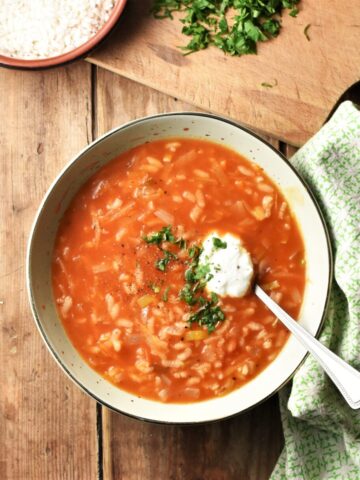 Tomato rice soup with yogurt and herbs in green bowl with spoon, green cloth the the right and rice in dish on top of wooden board with chopped herbs in background.