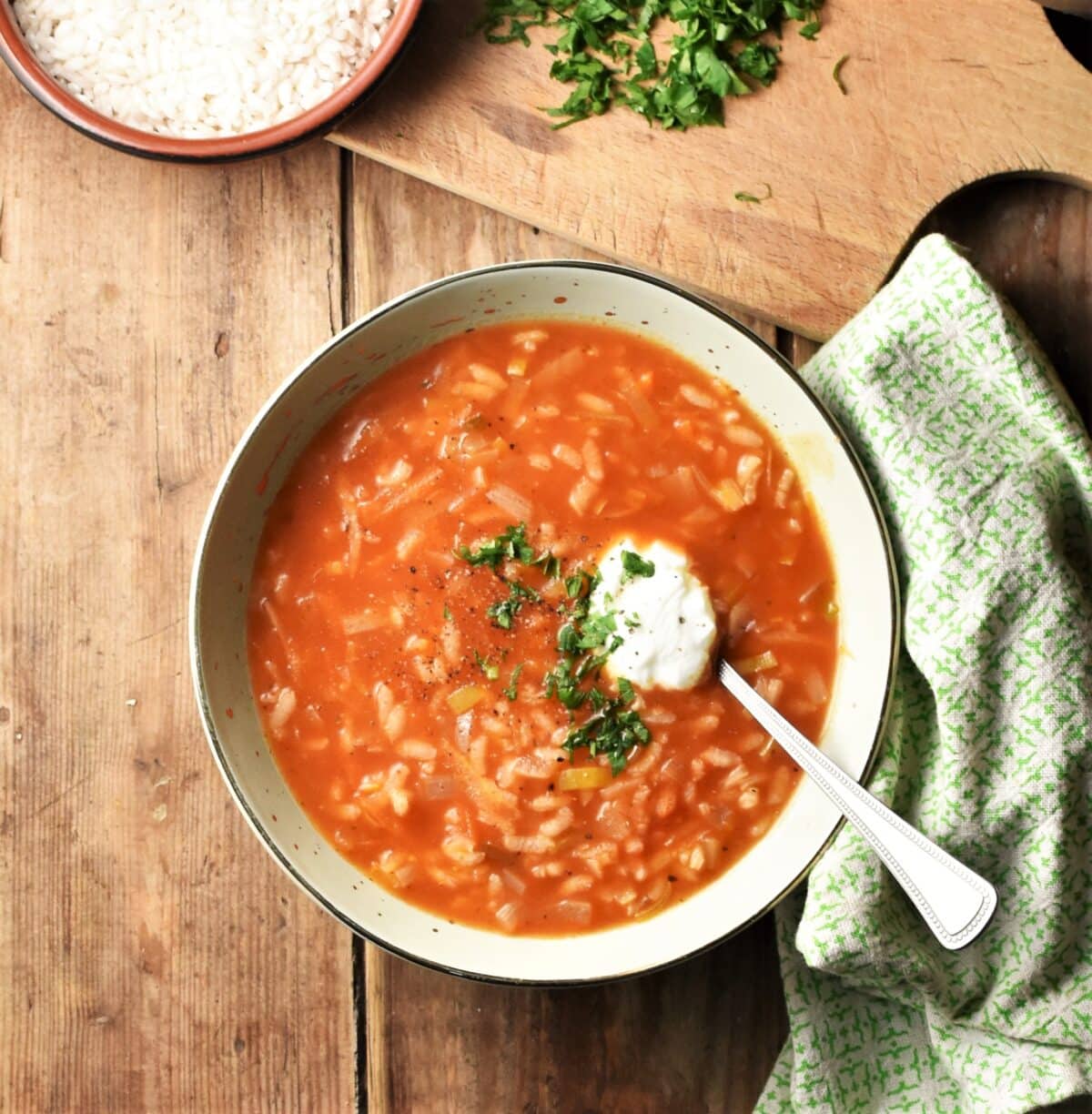 Tomato rice soup with yogurt and herbs in green bowl with spoon, green cloth the the right and rice in dish on top of wooden board with chopped herbs in background.