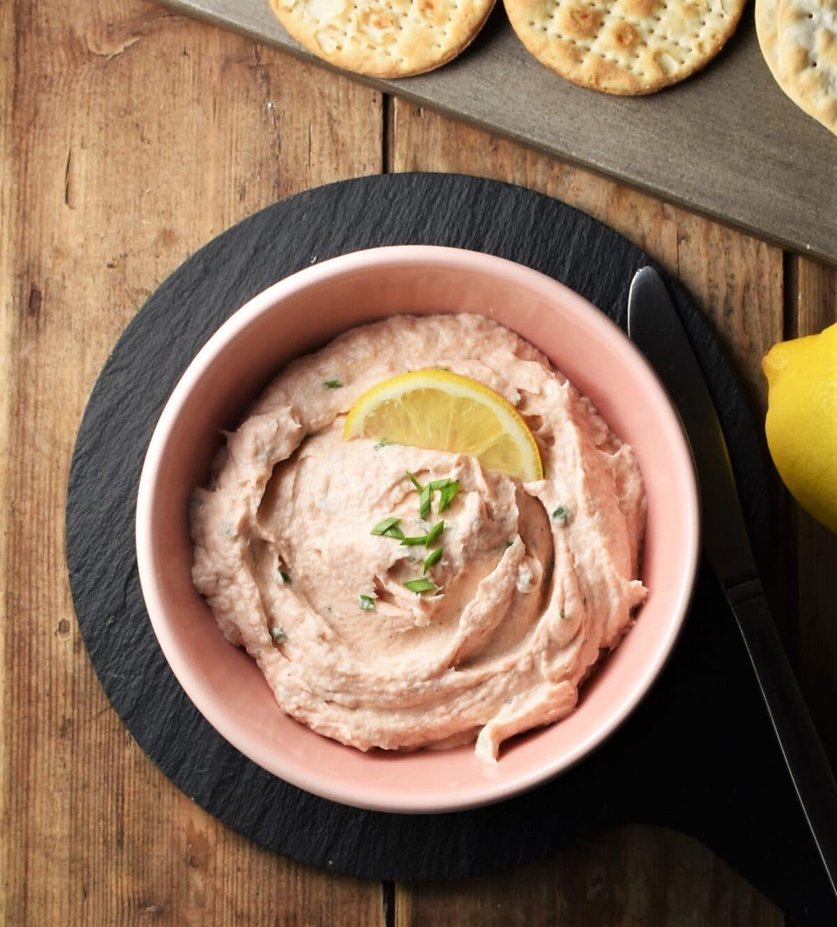 Creamy salmon spread with lemon slice in pink bowl on black plate with crackers and lemon in background.