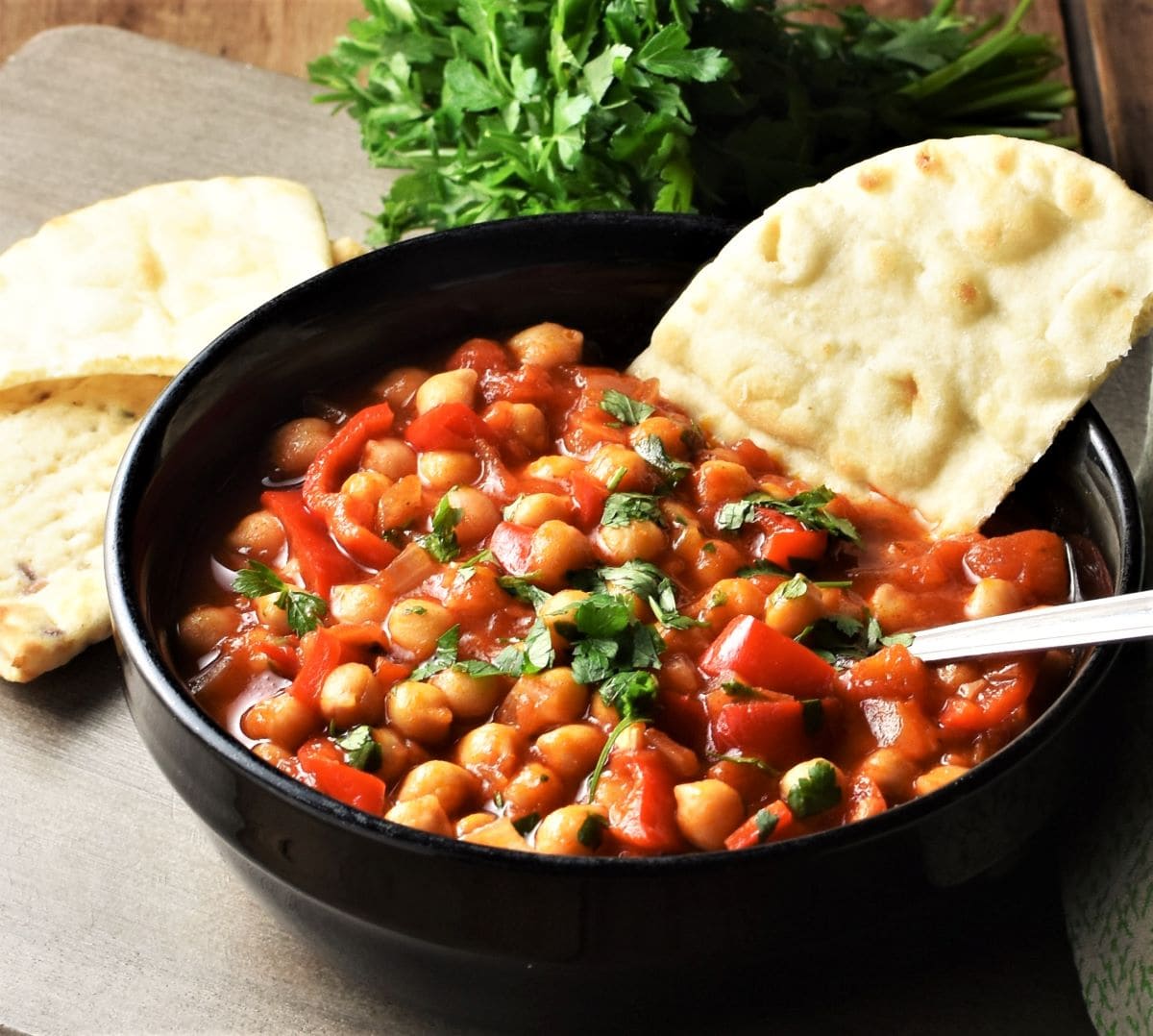 Side view of chickpea stew in black bowl with pita and parsley in background.
