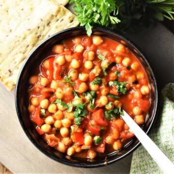 Chickpea stew in black bowl with spoon, fresh parsley and pita in background.