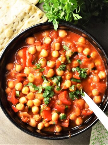 Chickpea stew in black bowl with spoon, fresh parsley and pita in background.