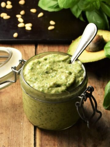 Side view of avocado pesto in jar with spoon, avocado, basil and pine nuts in background.