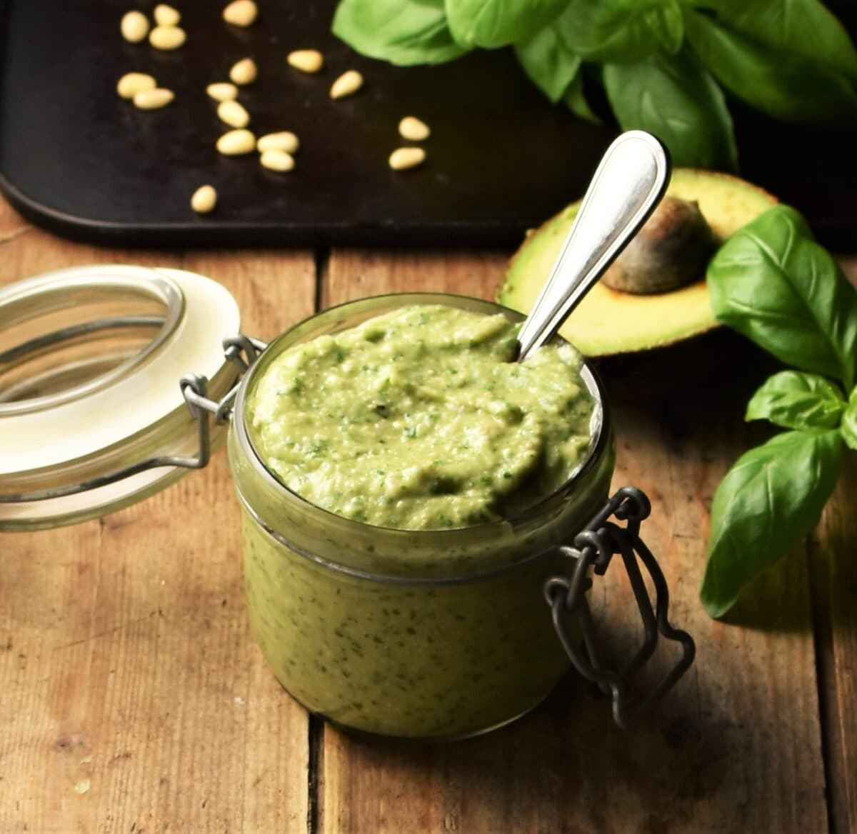 Side view of avocado pesto in jar with spoon, avocado, basil and pine nuts in background.