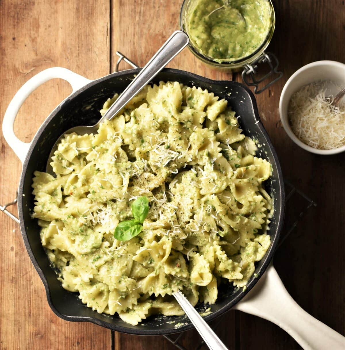 Top down view of avocado pesto pasta in skillet with 2 spoons, pesto and grated cheese in background.