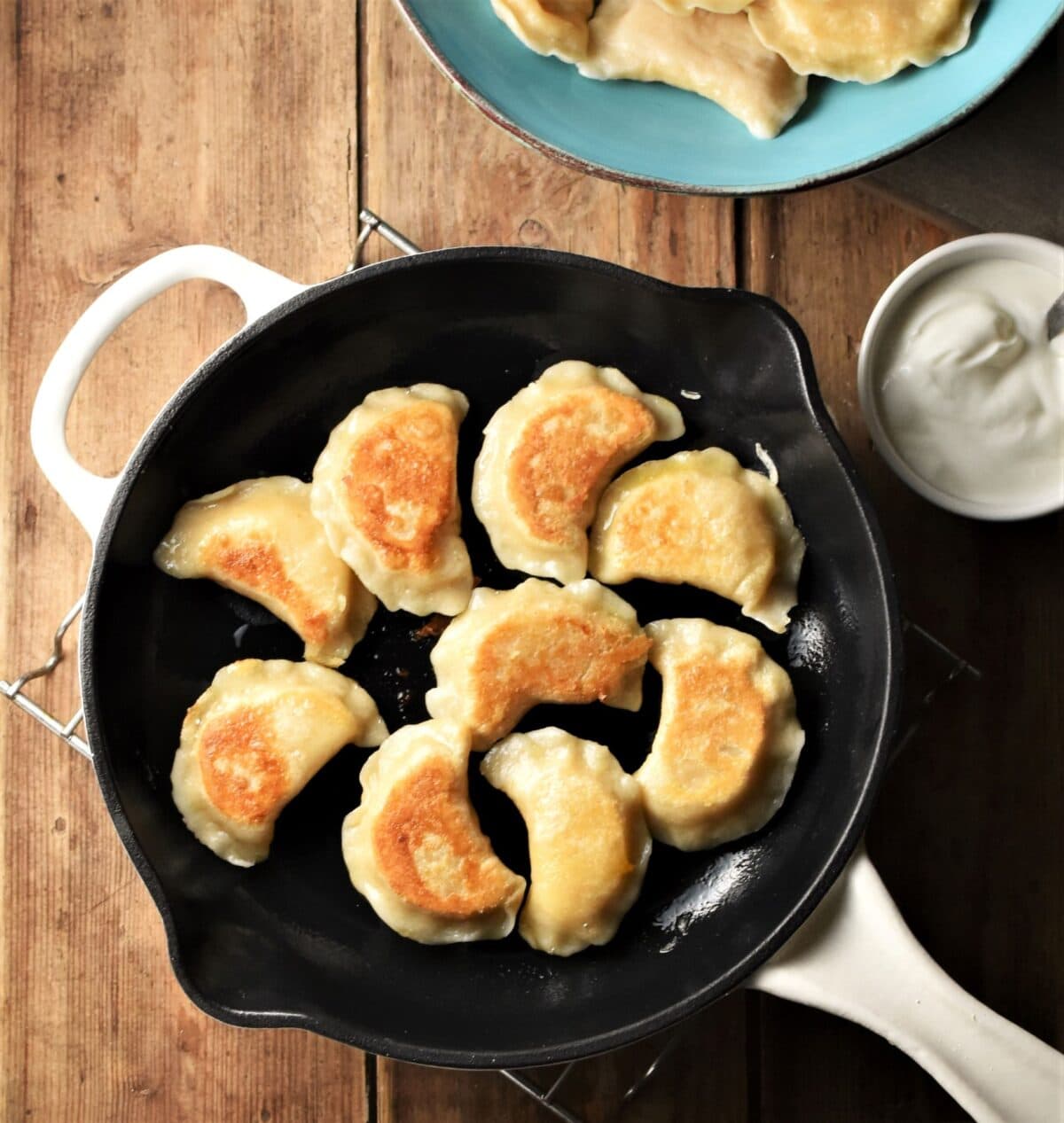 Fried pierogies in pan with sour cream and pierogies in blue bowl in background.